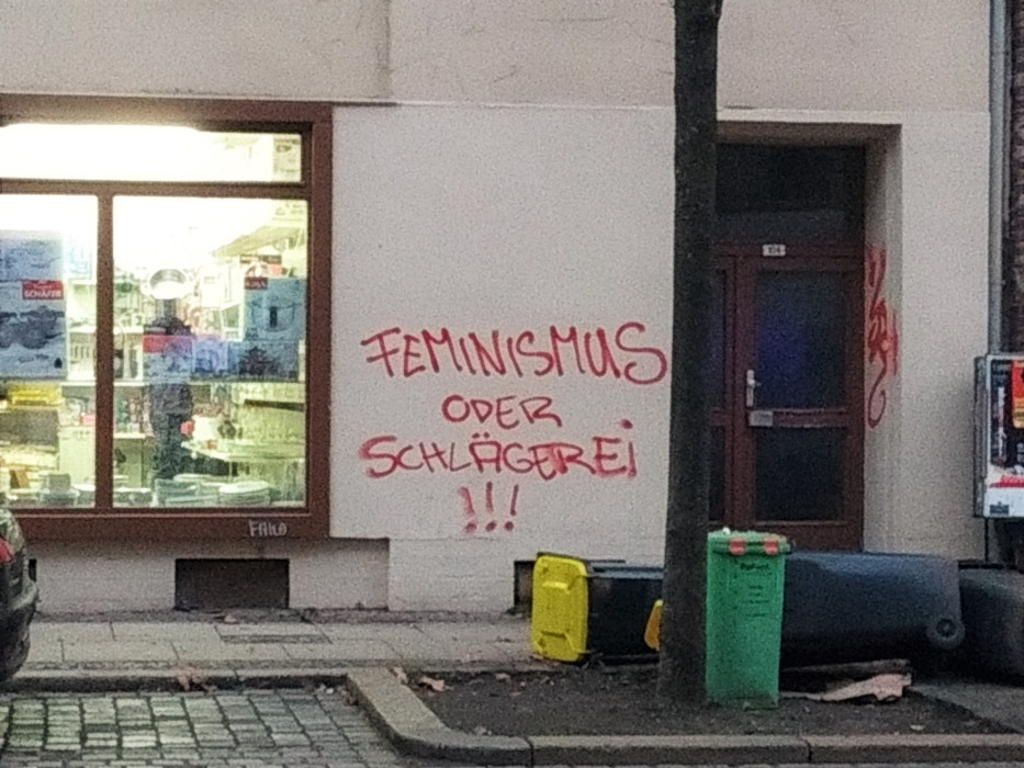 Straßenszene Eisenbahnstraße: Licht scheint durch ein Schaufenster, umgestoßene Mülltonnen und an der Hauswand steht "Feminismus oder Schlägerei!!!"